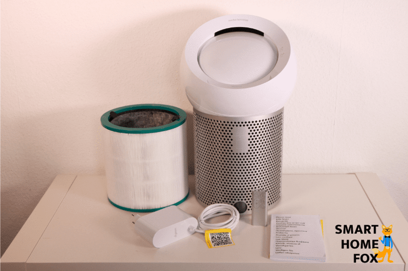 Der Dyson Pure Cool Me BP01 Luftreiniger und Tischventilator steht auf dem Regal. Daneben befindet sich die Glas-HEPA- und Aktivkohlefilter-Einheit, die Fernbedienung, das Stromkabel sowie die Bedienungsanleitung.